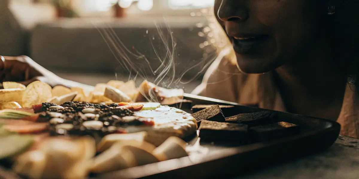 Close-up of a person smiling and inhaling steam from a food platter with cheese, crackers, and black olives, in soft indoor lighting.