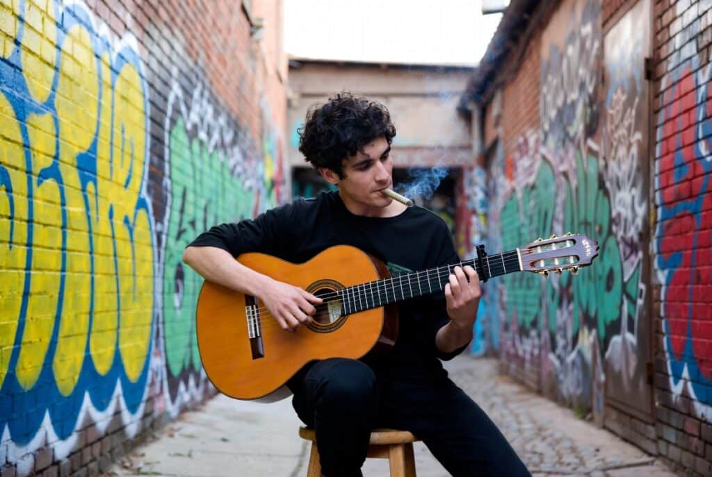 Top pre roll reviews street guitar smoker playing acoustic guitar in graffiti alley with lit pre-roll.