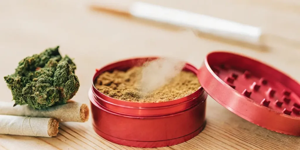 Close-up of a red grinder filled with cannabis kief next to rolled joints and a cannabis bud.