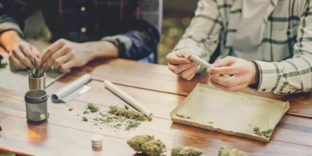Sophisticated scene with two people discussing cannabis, surrounded by neatly rolled joints, buds, and smoking accessories on a wooden table