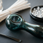 Clean cannabis smoking pipe tools with a glass pipe, cotton swabs, grinder, and scraper on a dark surface.