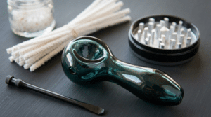 Clean cannabis smoking pipe tools with a glass pipe, cotton swabs, grinder, and scraper on a dark surface.