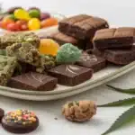 Variety of colorful gummies, brownies, cookies, and chocolates on a white plate, decorated with fresh cannabis leaves on a light surface