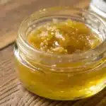 Close-up of CBD Sugar Wax in a jar with natural lighting and cannabis leaves on a wooden surface.