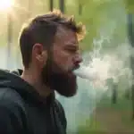 Bearded man exhaling cannabis smoke in the forest, potentially suffering from burnt throat.