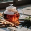 Darker cannabis root tea in a clear teapot, with cannabis roots and leaves arranged nearby.
