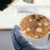A gloved hand examining a petri dish with microorganisms under a microscope.