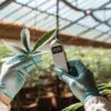 Gloved hands holding a cannabis leaf and a scientific device in a bright greenhouse.