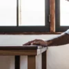 Close-up of a man's hand gripping a wooden table edge, next to a sunlit window.