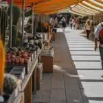 Wide-angle shot of a bustling open-air market with stalls of cactus and other plants, under orange and white striped awnings, with blurred people walking.