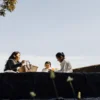 Family enjoying a peaceful outdoor picnic on a sunny day with trees and open sky surrounding them.