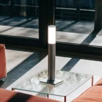 Modern minimalist table lamp glowing on a glass coffee table in a bright living room.