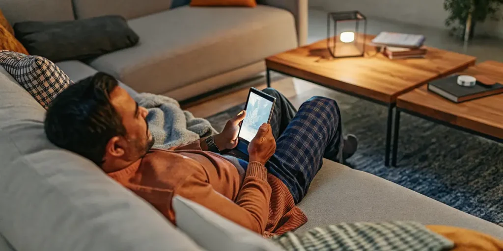 Man relaxing on a sofa in a cozy living room while using a tablet under warm ambient lighting.