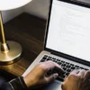 Close-up of hands typing programming code on a laptop beside a desk lamp.