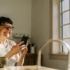 Smiling middle-aged woman using her smartphone while sitting at a bright kitchen table.