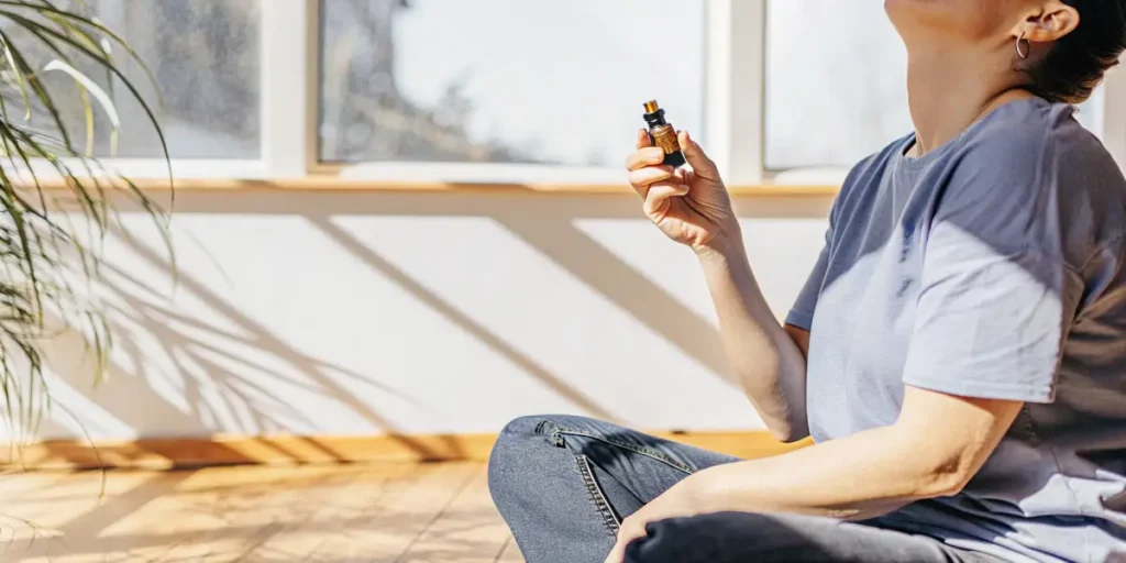 Person sitting on the floor holding a CBD oil bottle during a relaxing aromatherapy session by a sunlit window.