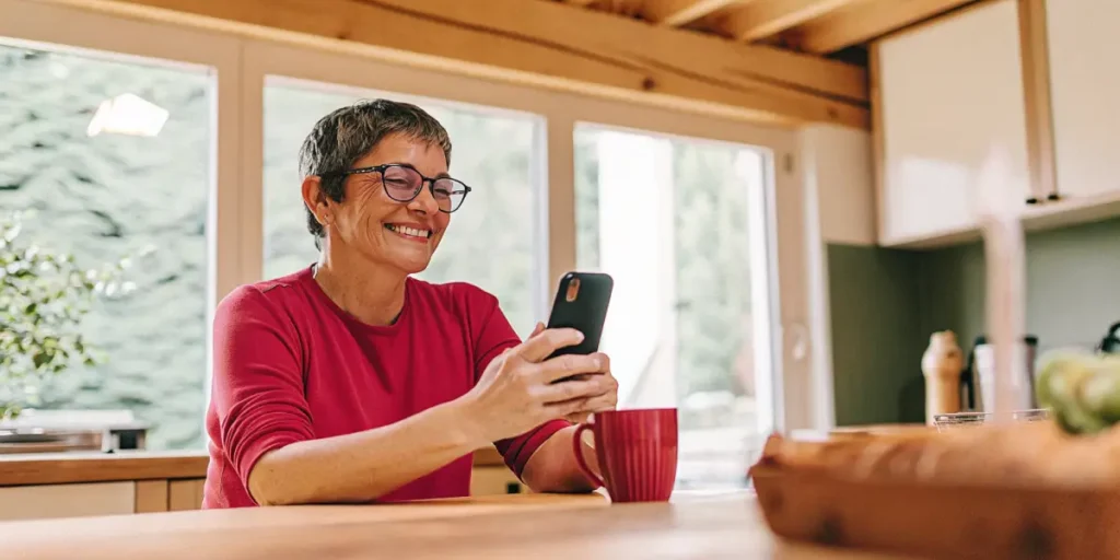 Smiling woman checking her smartphone while holding a coffee cup in a cozy kitchen.