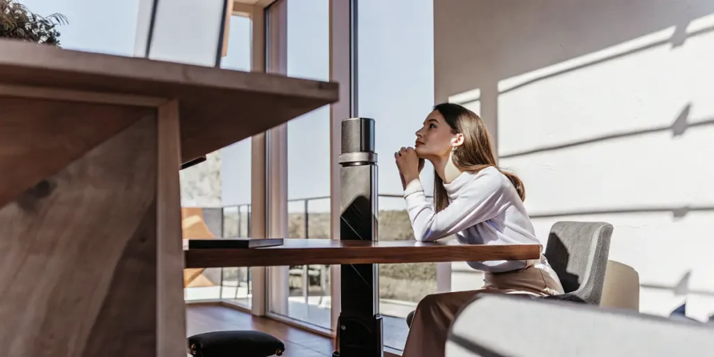 Thoughtful woman sitting at a modern desk, looking up with her hands under her chin in a bright workspace.