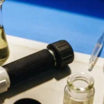 Handheld refractometer placed on a laboratory bench beside cannabis samples and glass flasks during brix analysis.
