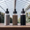 Three bottles filled with cannabis terpene powders displayed on a wooden table inside a greenhouse surrounded by flowering cannabis plants.