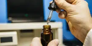 Hand using a dropper to dispense cannabinoid oil into a glass bottle in a laboratory setting.