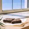 Cannabis brownie edible placed on parchment paper near a window, surrounded by cannabis leaves in natural light.