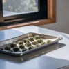 Cannabis buds evenly spaced on a metal baking tray lined with parchment paper, placed on a kitchen counter near a sunlit window.