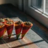 Colorful cannabis candy cones filled with ground marijuana buds arranged on a wooden table near a window.