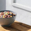 Cannabis-infused colorful cereal mix in a ceramic bowl placed on a wooden table near a sunlit window.