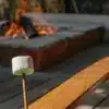 Cannabis-infused marshmallow on a wooden stick roasting near an outdoor fire pit at sunset