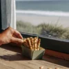 Hand reaching for crispy cannabis seed crackers in a small box placed on a wooden windowsill with ocean view in the background.