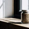 Glass jar filled with cannabis seeds stored on a wooden shelf near a sunlit window.