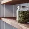 Glass jar filled with cured cannabis buds displayed on a wooden shelf near a window.