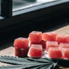 Red THC gummies arranged on a white plate beside a cannabis leaf on a wooden table near a window with natural sunlight.