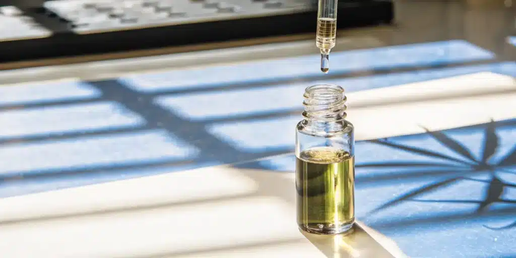 Cannabis oil dropper above glass vial with golden extract in natural sunlight.