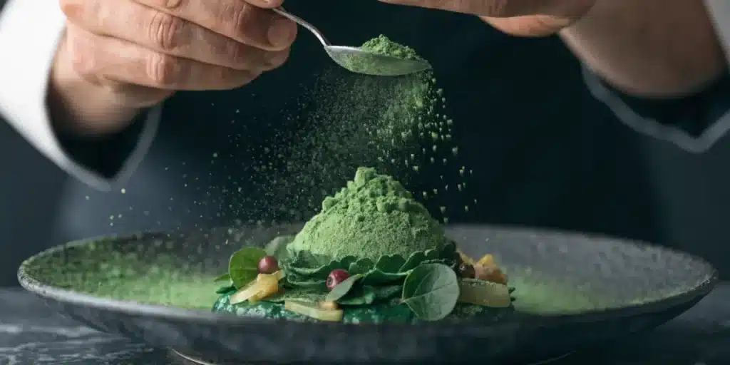 Chef adding cannabis oil powder to a plated gourmet dish with green ingredients.
