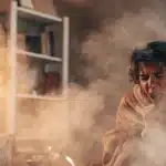 Person coughing in a smoky room after inhaling cannabis smoke.