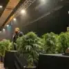 A presenter addresses the audience as premium cannabis plants are displayed next to a championship trophy during a live cannabis cup competition. The event showcases top genetics while judges and spectators observe the entries.