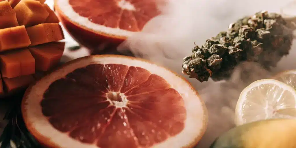 Macro close-up of grapefruit slices and cannabis buds with visible aromatic vapor and tropical fruit accents.