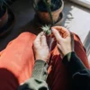 Person examining cannabis bud near window with indoor plants and sunlight.